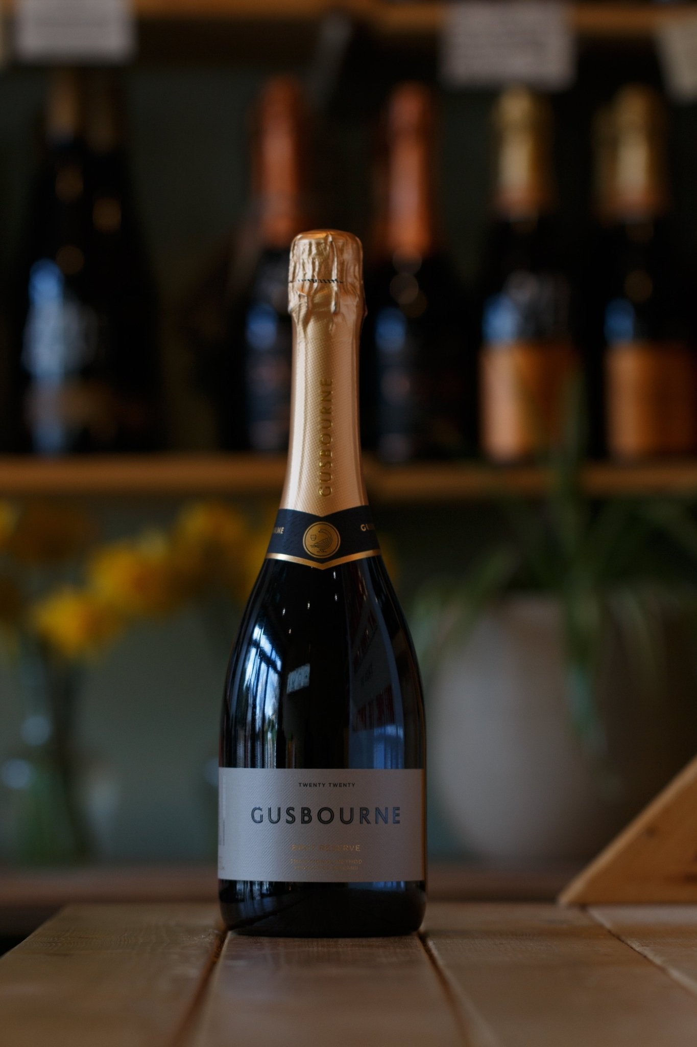 Gusbourne Brut Reserve 2020 sparkling wine from Pulp, displayed elegantly on a wooden table with a backdrop of various wine bottles, showcasing freshness and sophistication for any celebration.
