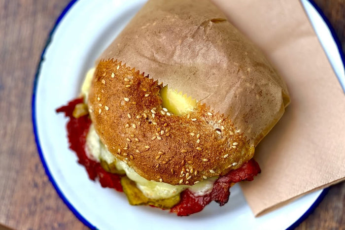 Sandwich on a sesame seed bun with visible ingredients on a white plate with blue rim, placed on a wooden surface.