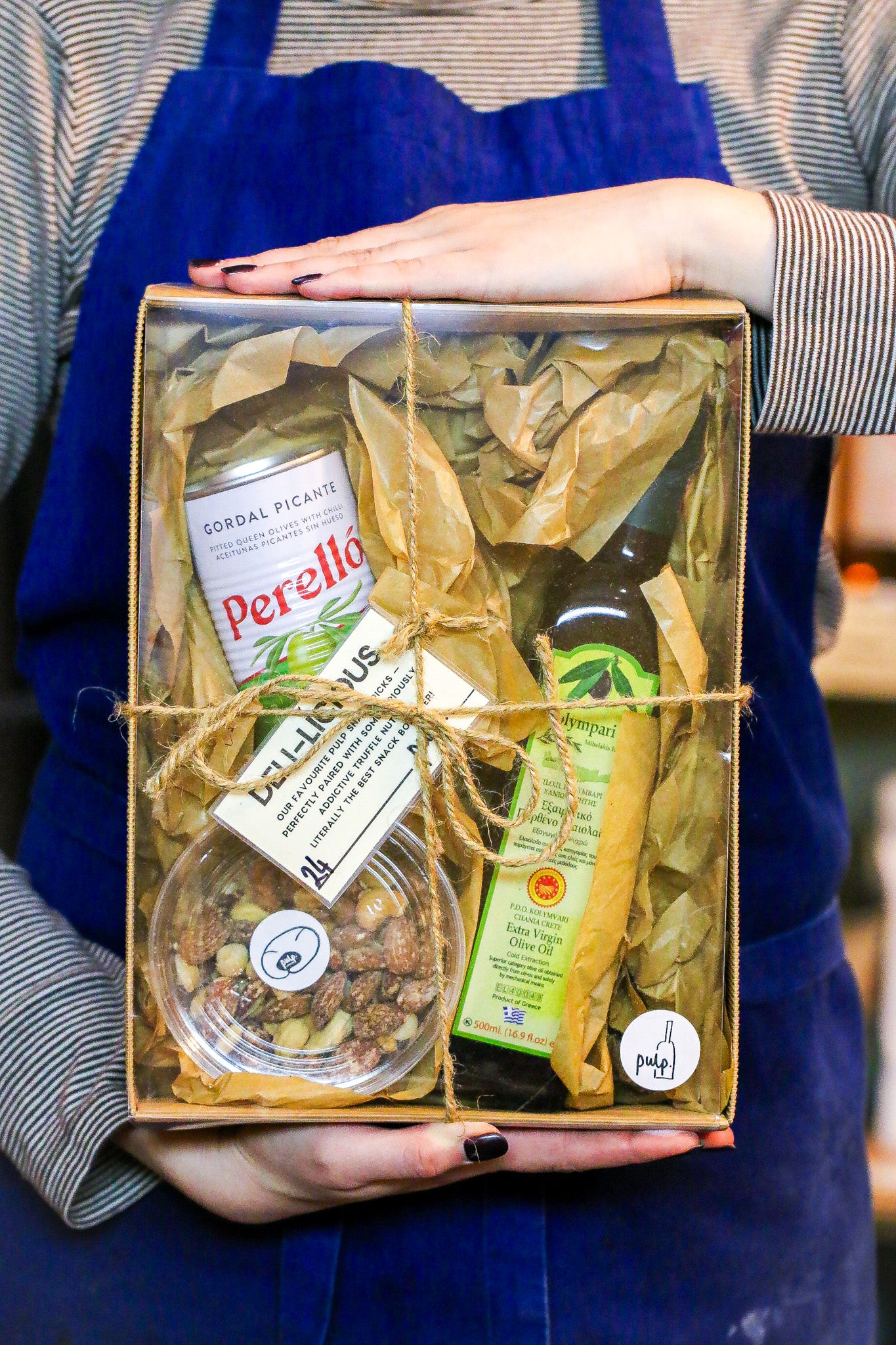 Person holding a gift box with food items including a bottle of olive oil and a jar of olives.