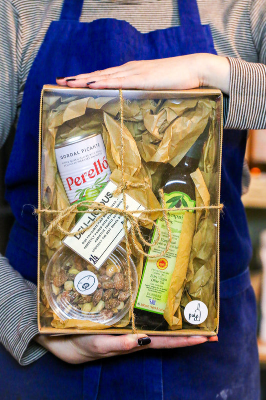 Person holding a gift box with food items including a bottle of olive oil and a jar of olives.