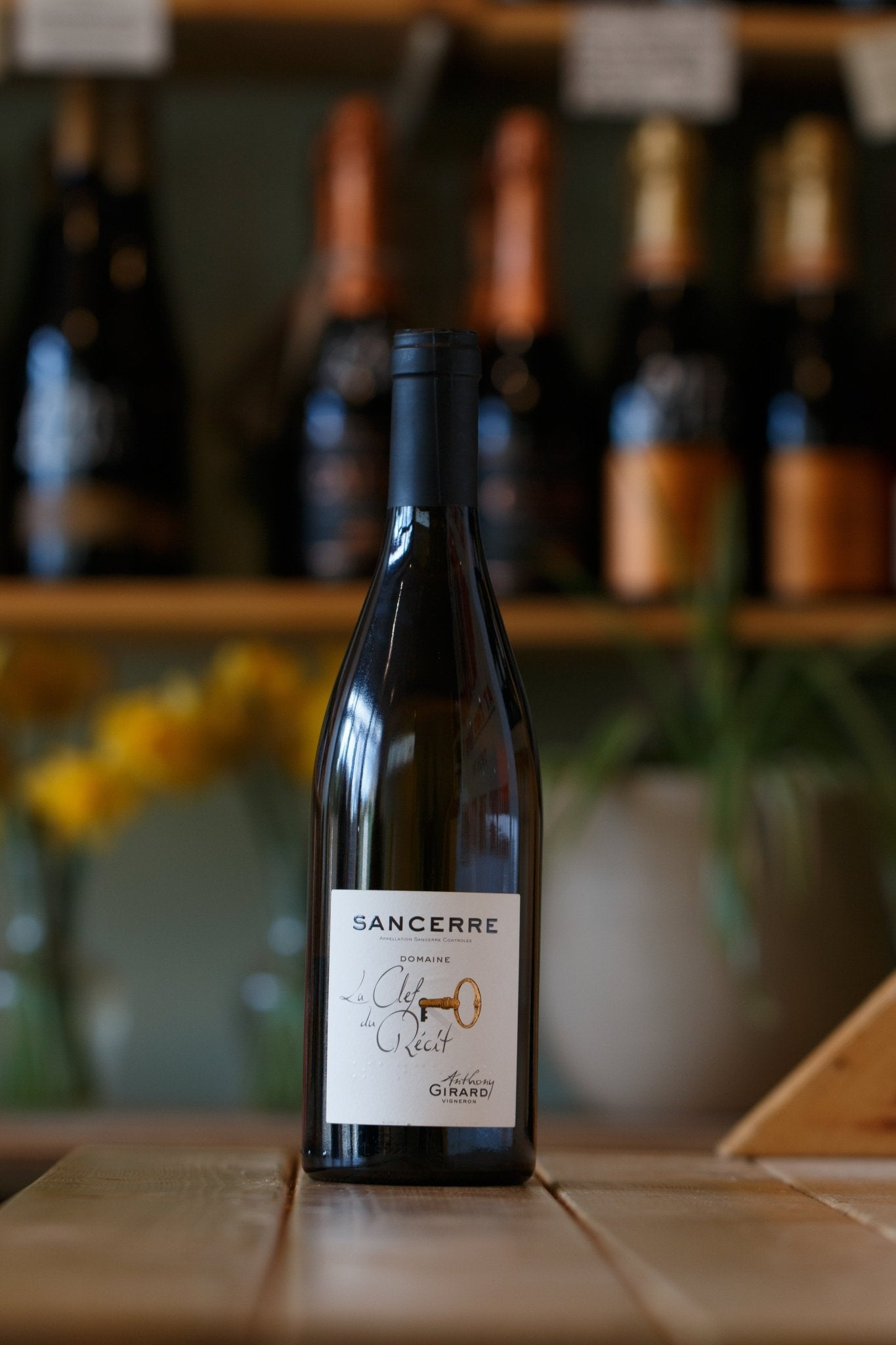 La Clef Du Récit Sancerre 2023 by Anthony Girard displayed elegantly on a wooden table, with a background of blurred wine bottles, showcasing the premium organic Sauvignon Blanc from the Loire Valley, France.