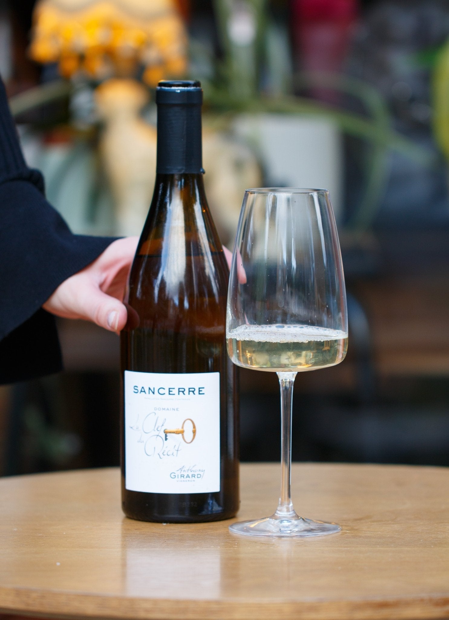 A person holding a glass next to a bottle of La Clef Du Récit Sancerre 2023 by Anthony Girard, highlighting the elegant organic Sauvignon Blanc, perfect for any occasion.